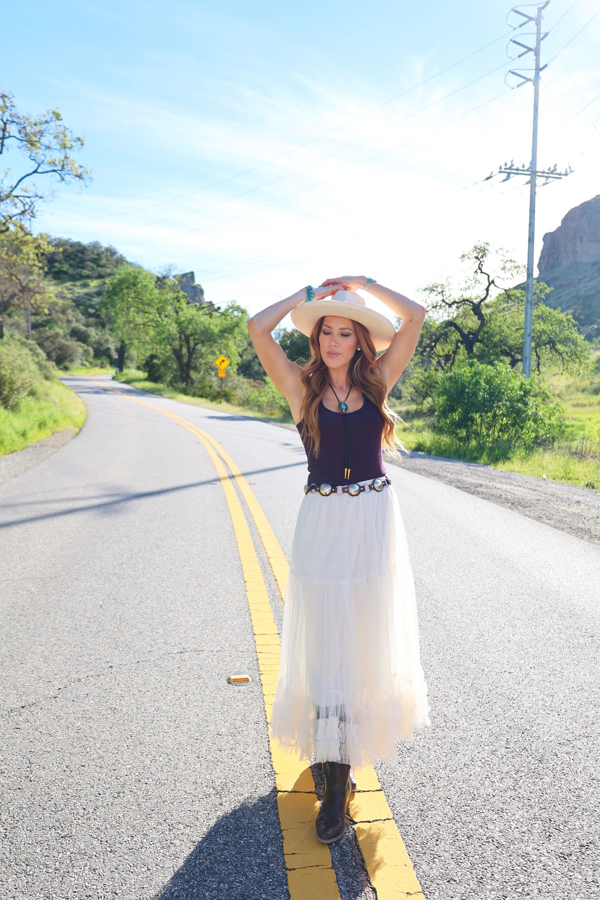 Kimberly Dawn in a cream hat, dark tank top and white tulle skirt on a winding country road, from the Highway Heartache shoot