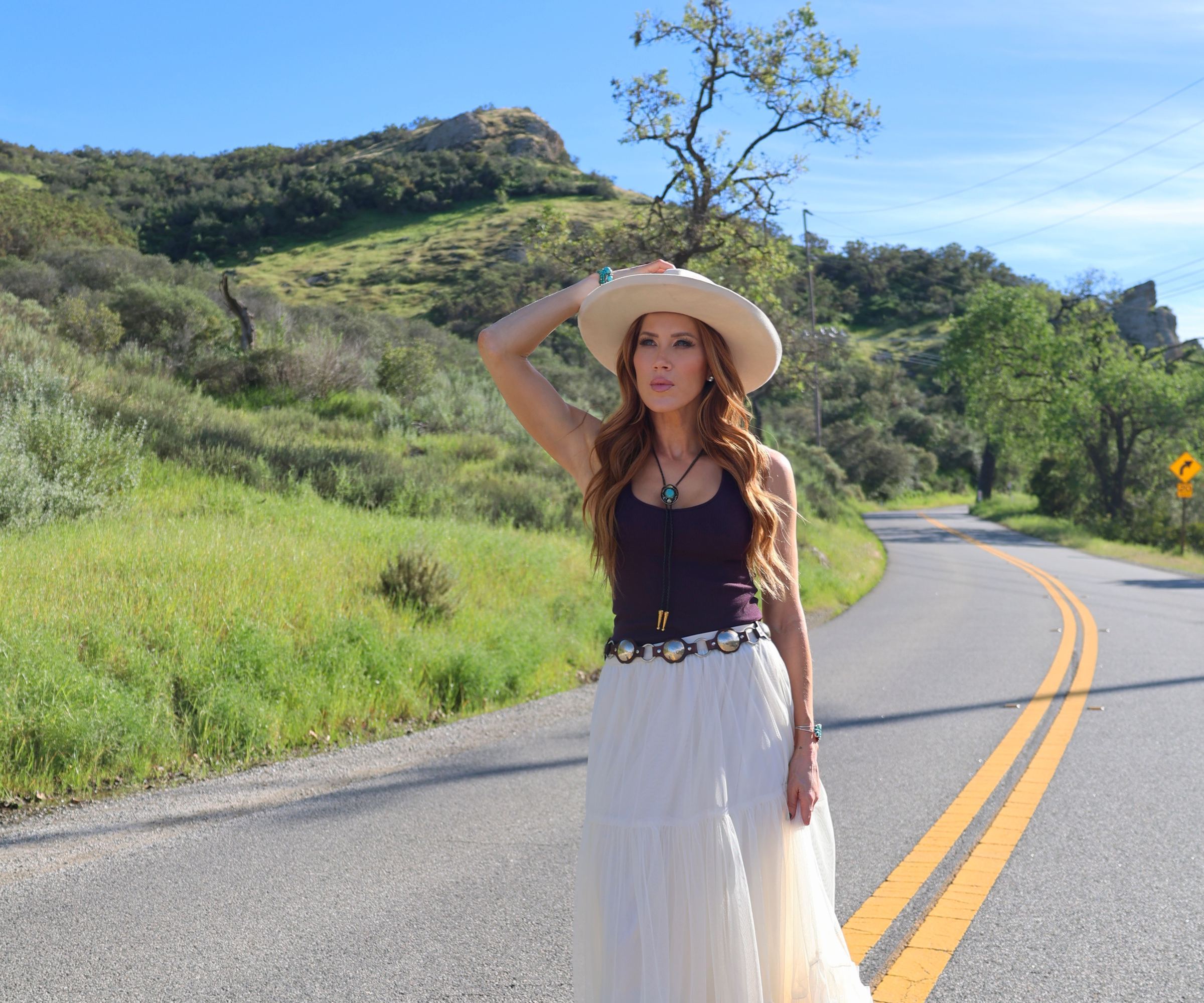 Kimberly Dawn in a cream hat, dark tank top and white tulle skirt on a winding country road, from the Highway Heartache shoot